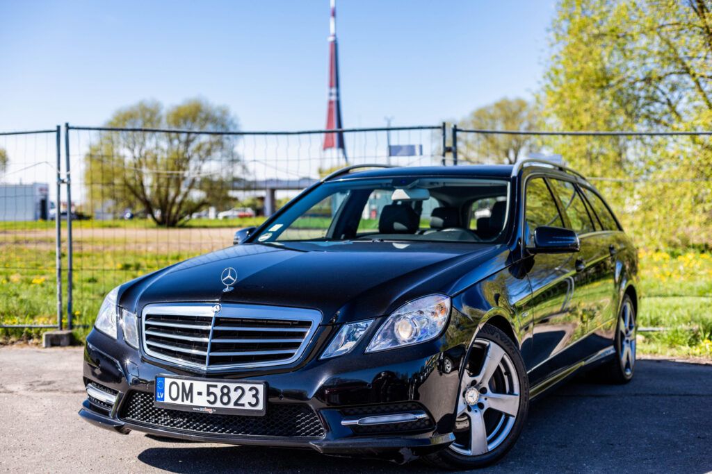 Front view of a black Mercedes SUV with license plate OM-5823 in a sunny parking lot, fence and a tall rocket structure in the background.
